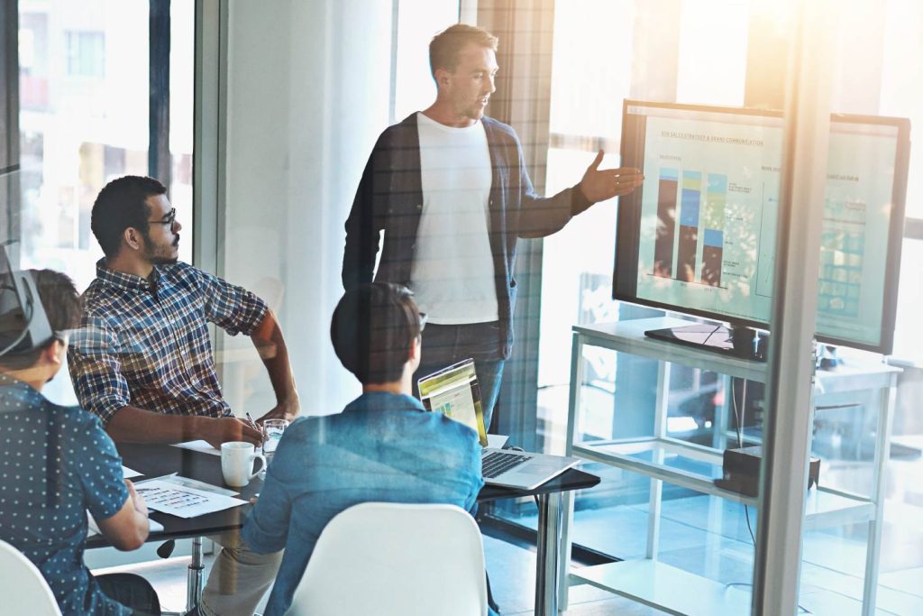 Team in a meeting room with the presenter pointing at data report graphs on a large monitor