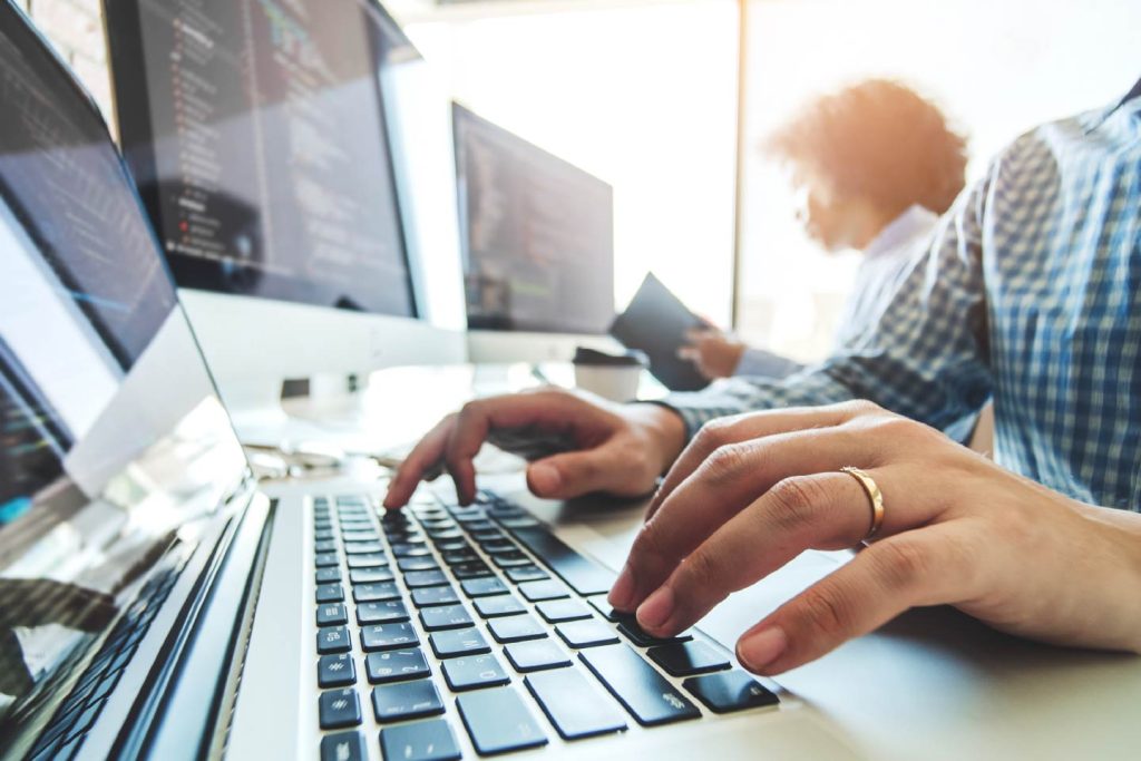 Close up of hands typing on a keyboard with multiple computer monitors and a work colleague in the background
